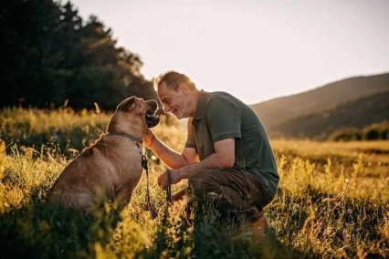 Profile view of senior man kneeling and smiling at his dog and petting the dog. They are in a meadow with tall hills in the background.