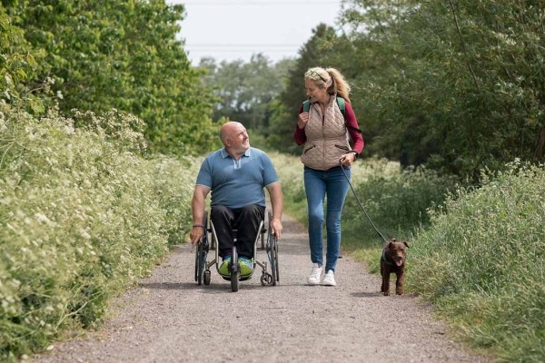 Un hombre con paraplejía en silla de ruedas y una mujer caminando, en un día soleado de verano en una reserva natural con su perro.